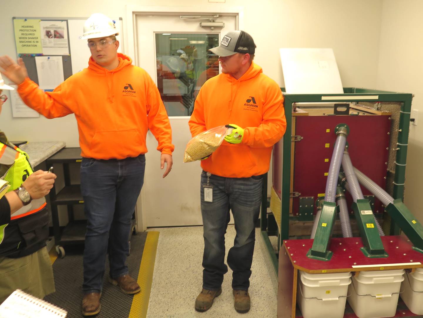 Hunter Girten (left), Bayer Commercial Soybean Processing Facility production associate, explains the role of the shaker machine (right) located in the quality lab, as Reid Mathenia, seed technician, holds a bag of soybeans that went through the process that eliminates debris from the seed soybeans before they are tested in the lab.