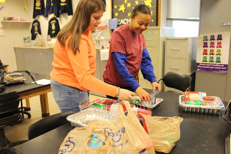 Ella Chalkey (left) and Kiana Hawthorne demonstrate how the Cake in a Pan kits will be assembled by Streator FFA members at the chapter’s Lock-In during National FFA Week. Once the kits are completed, FFA members will deliver them to the Streatorland Food Pantry for distribution to the community.
