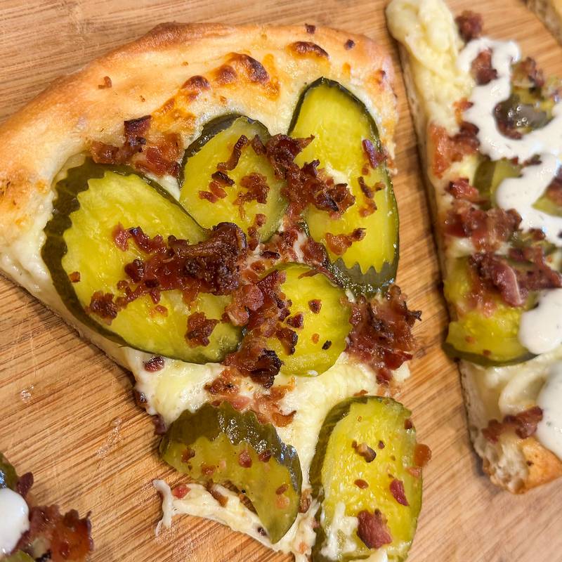 Pickle Bacon Ranch Pizza by Swains Concessions. A homemade pizza dough is stretched to perfection and covered with an award-winning pickle ranch sauce and mozzarella cheese. Topped off with pickles and bacon, with a touch of pickle ranch to finish it off.
