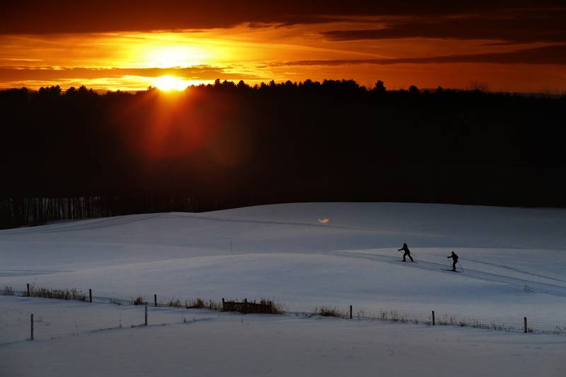 A pair of cross-country skiers race over snow-covered hills as the sun sets on a fine winter day.